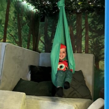 a small child in a pumpkin costume sits inside the sensory swing, smiling.