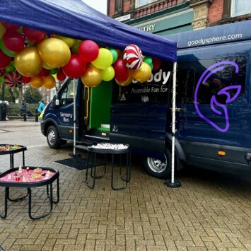 Image taken from an angle where the logo is clearly central on the rear of the bus, the sensory unit is open with three tuff trays outside under a purple gazebo.