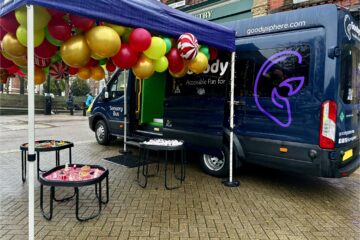 Image taken from an angle where the logo is clearly central on the rear of the bus, the sensory unit is open with three tuff trays outside under a purple gazebo.