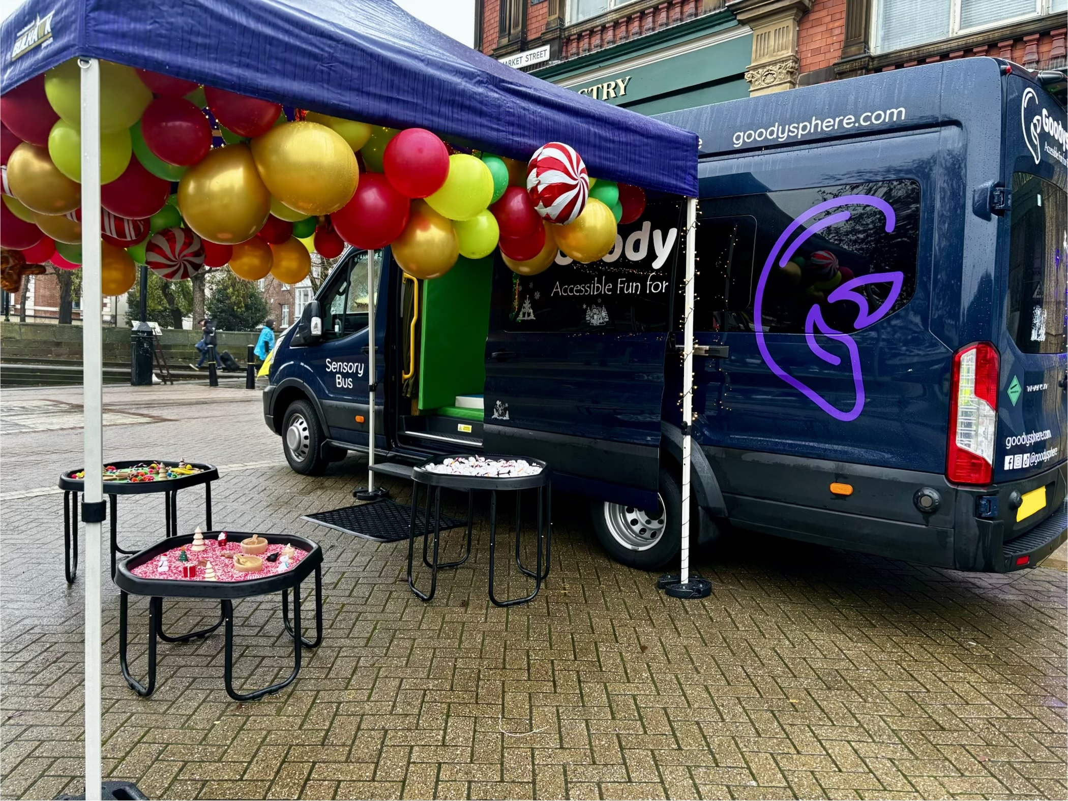 Image taken from an angle where the logo is clearly central on the rear of the bus, the sensory unit is open with three tuff trays outside under a purple gazebo.
