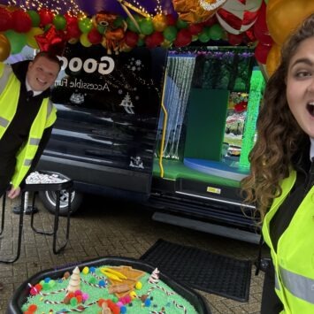 Two smiling staff members wearing high-visibility jackets smile in front of a sensory tray containing green rice and Christmas tree toys and the open sensory bus is visible in the background.