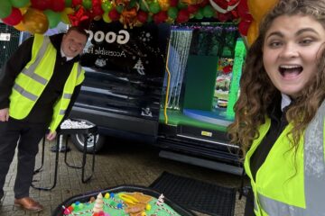 Two smiling staff members wearing high-visibility jackets smile in front of a sensory tray containing green rice and Christmas tree toys and the open sensory bus is visible in the background.