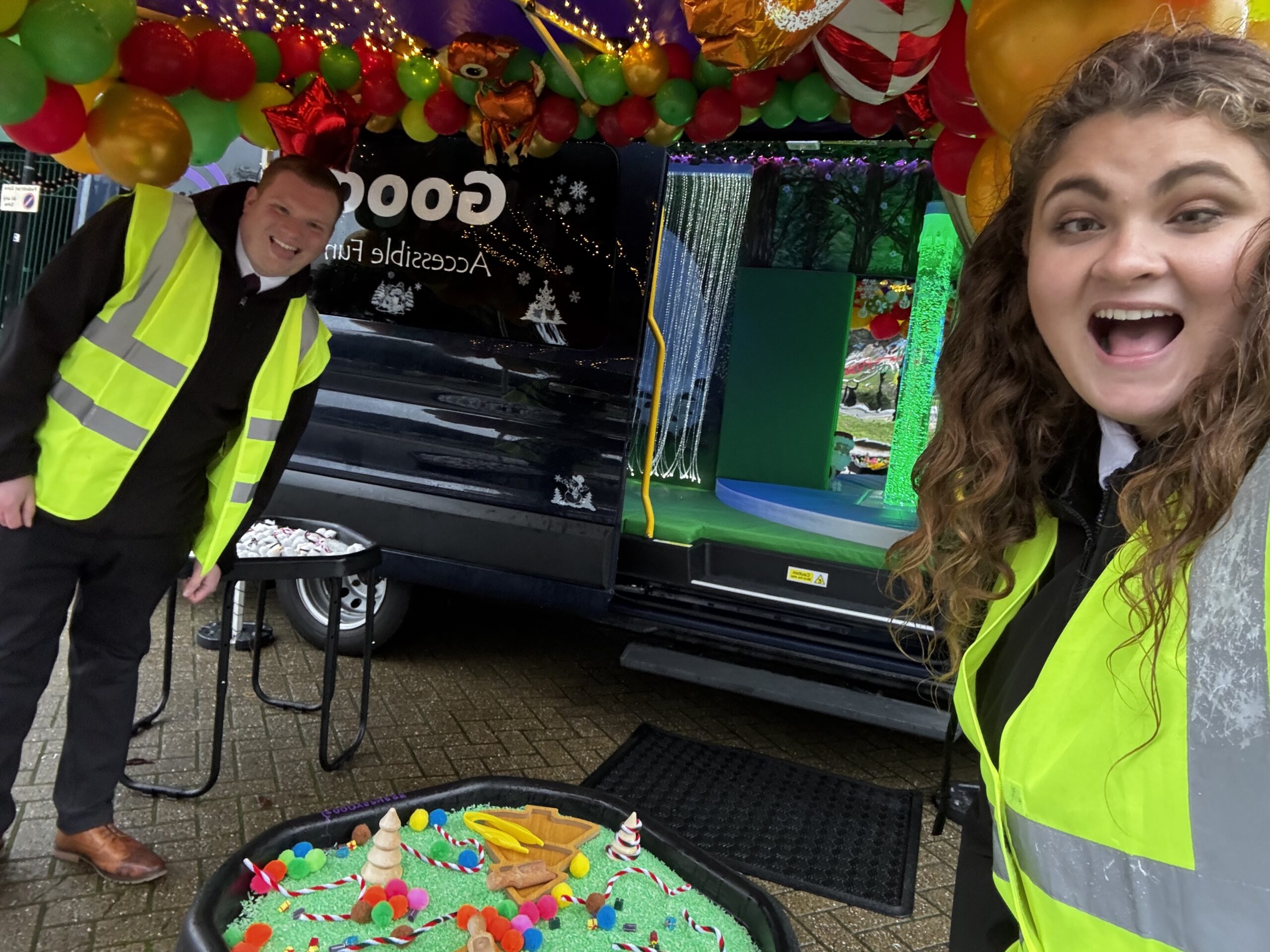 Two smiling staff members wearing high-visibility jackets smile in front of a sensory tray containing green rice and Christmas tree toys and the open sensory bus is visible in the background.