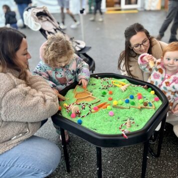 Two mums with their young children playing with a green Christmas tree sensory tray.