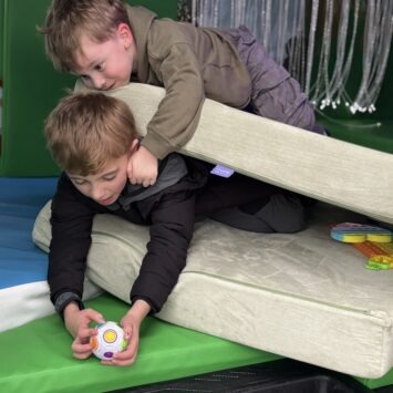 One boy is playing with a sensory toy whilst he is sandwiched in the play sofa and a younger boy lays on top.