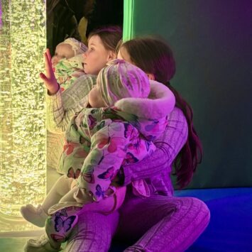 An older girl hold her baby sibling whilst interacting with the yellow bubbles on board the mobile sensory unit.