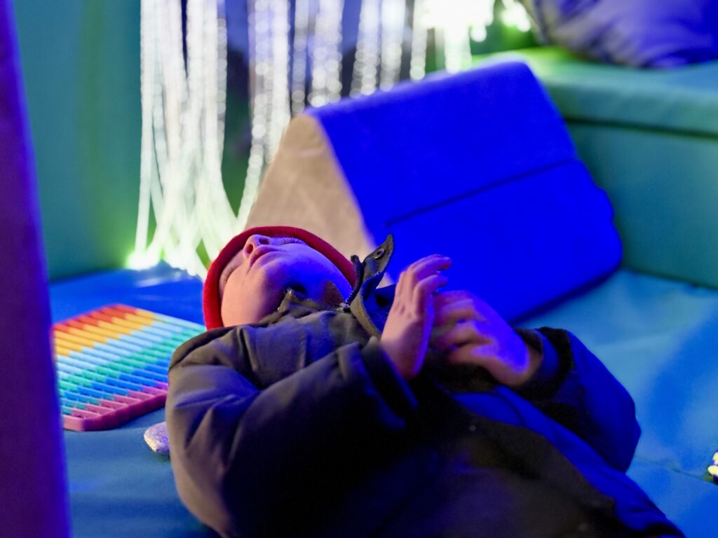 a young boy lays down looking up at the bubble tube, toys are scattered around him and the fibre optic curtain is blurred creatively behind him.