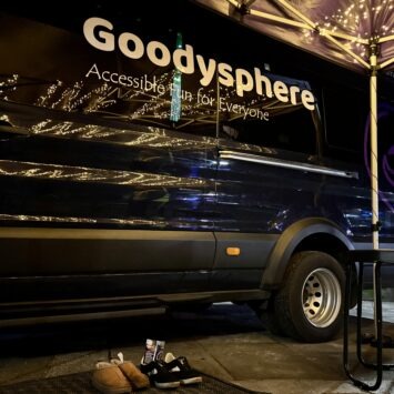 The door of the Goodysphere Sensory bus is closed reflecting the fairy lights on the gazebo. In front of it stand two pairs of shoes with a leaflet tucked in one shoe.
