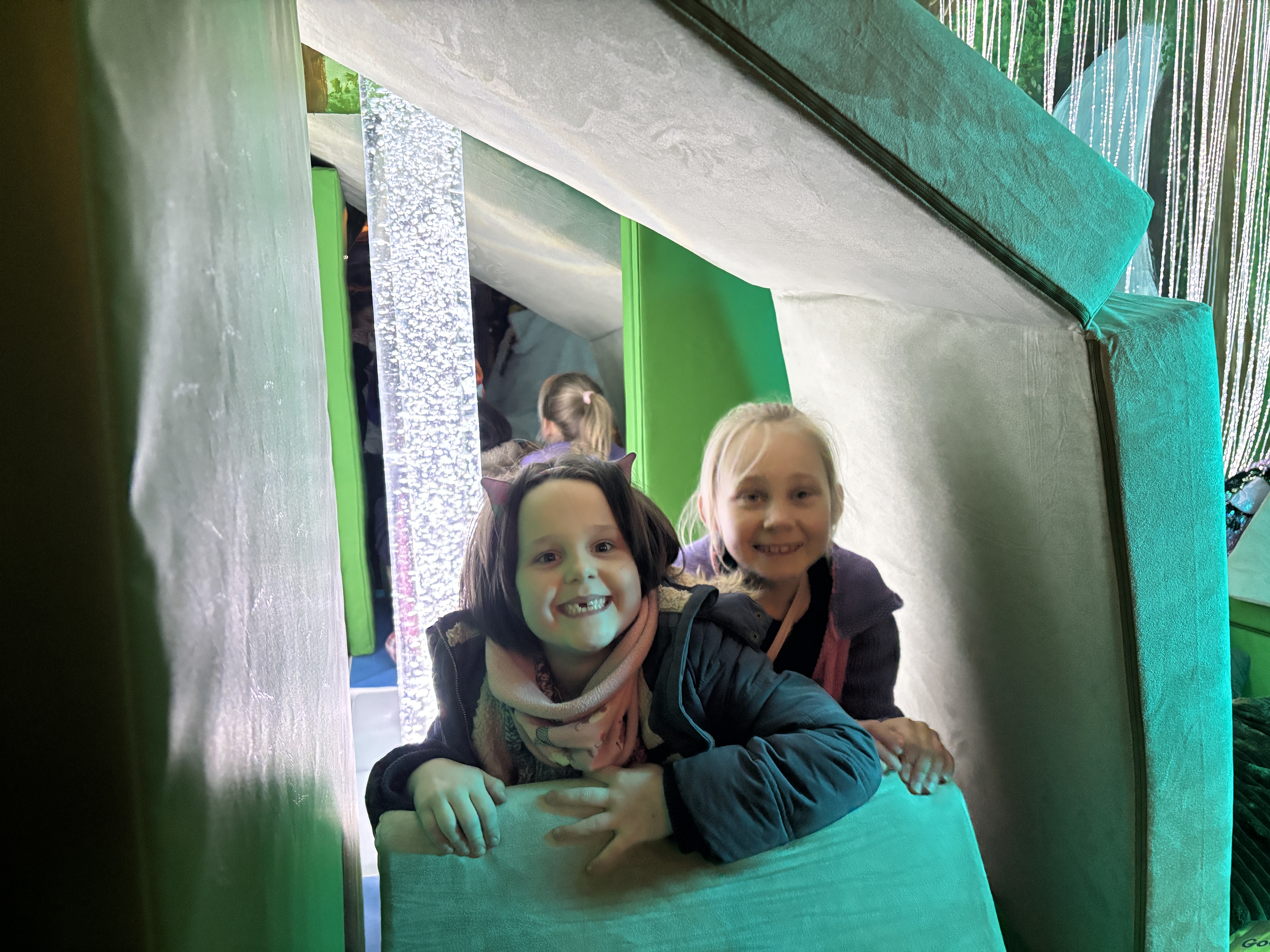 Two sister sit in a den they have built in front of a white bubble tube.