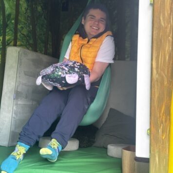 A young man smiles in the sensory swing whilst holding a sequin pillow