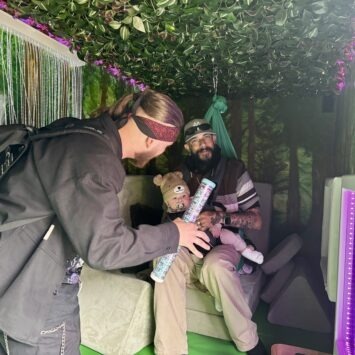 A Man wearing a baby in a slit, sits inside the sensory swing whilst his other adult friend engages with the rainmaker.